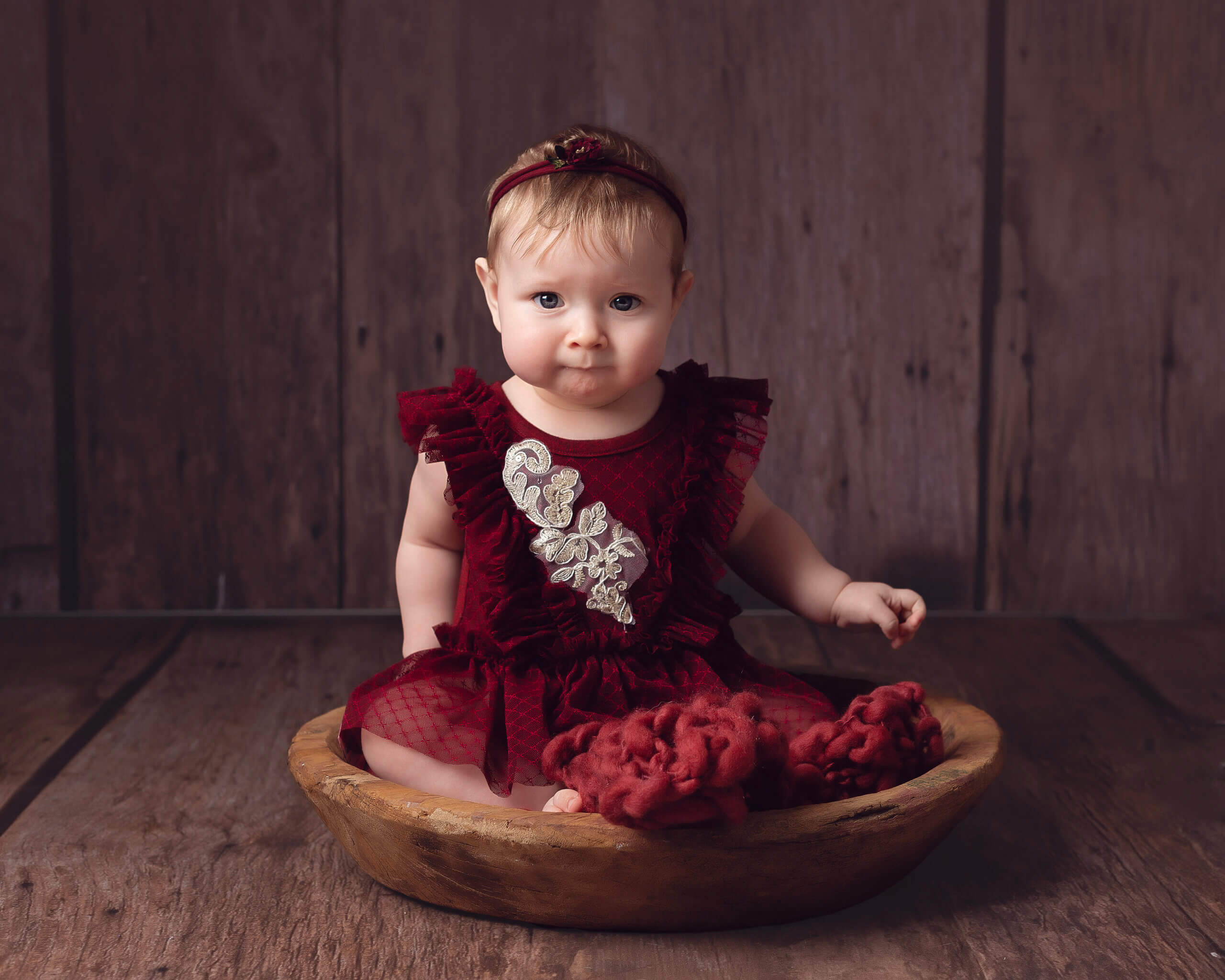 Cute baby girl in Maroon outfit sitting in a wooden bowl at her sitter photoshoot in Sutton Coldfield Birmingham